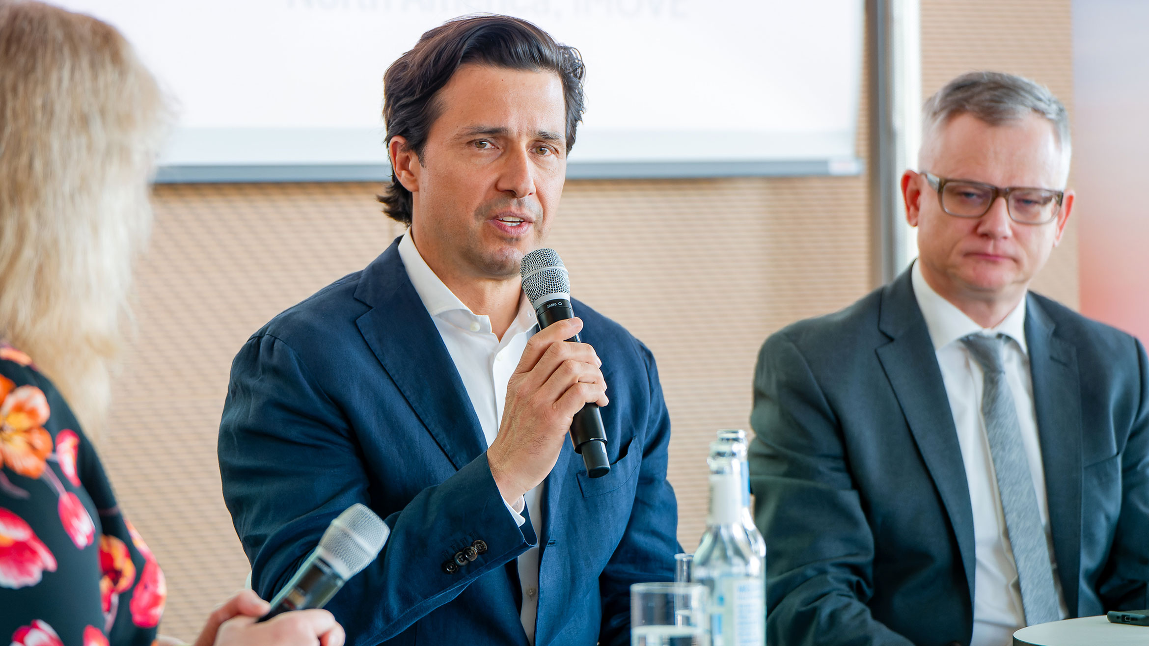 drei Menschen sitzen auf einem Podium, der Mann in der Mitte spricht in ein Mikrofon
