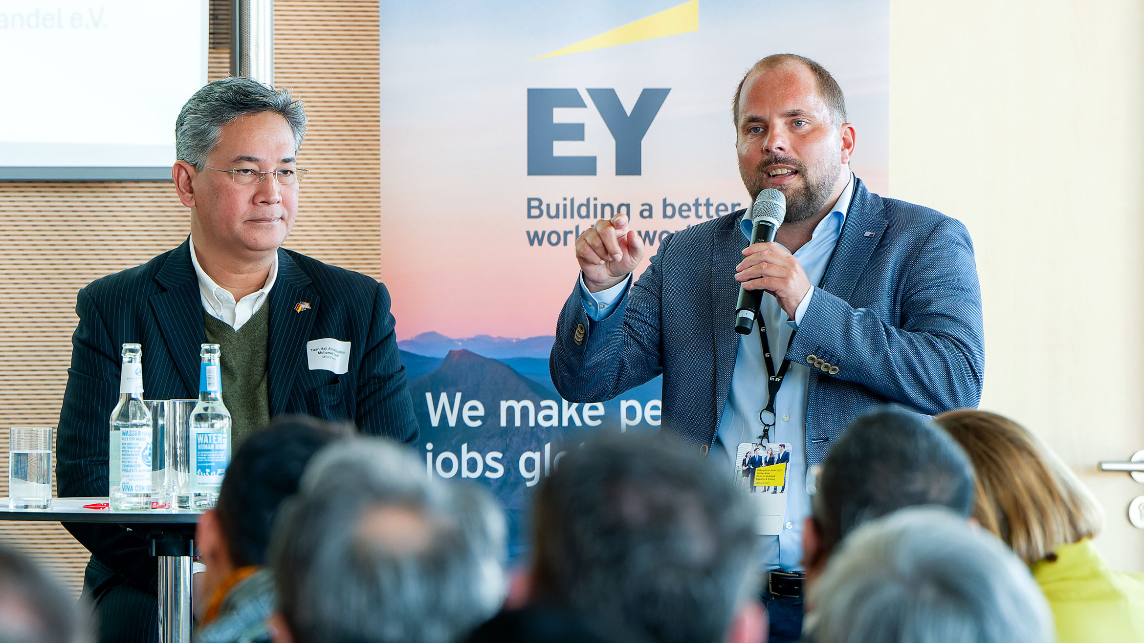 zwei Männer sitzen auf einem Podium, der Mann rechts spricht in ein Mikrofon