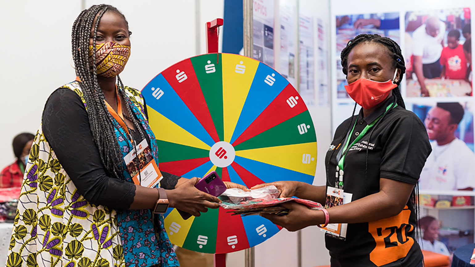 zwei Afrikanerinnen mit Mund-Nasen-Schutz stehen an Glücksrad mit Sparkassen-Logo