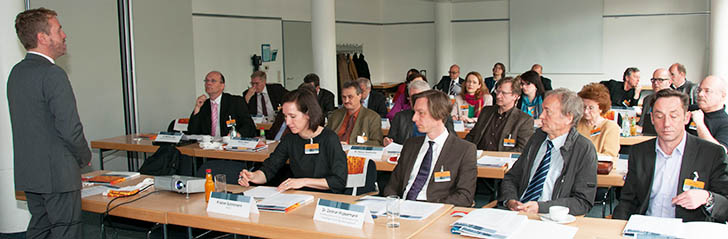 Blick auf alle Teilnehmer an Tischen und den Moderator davor stehend
