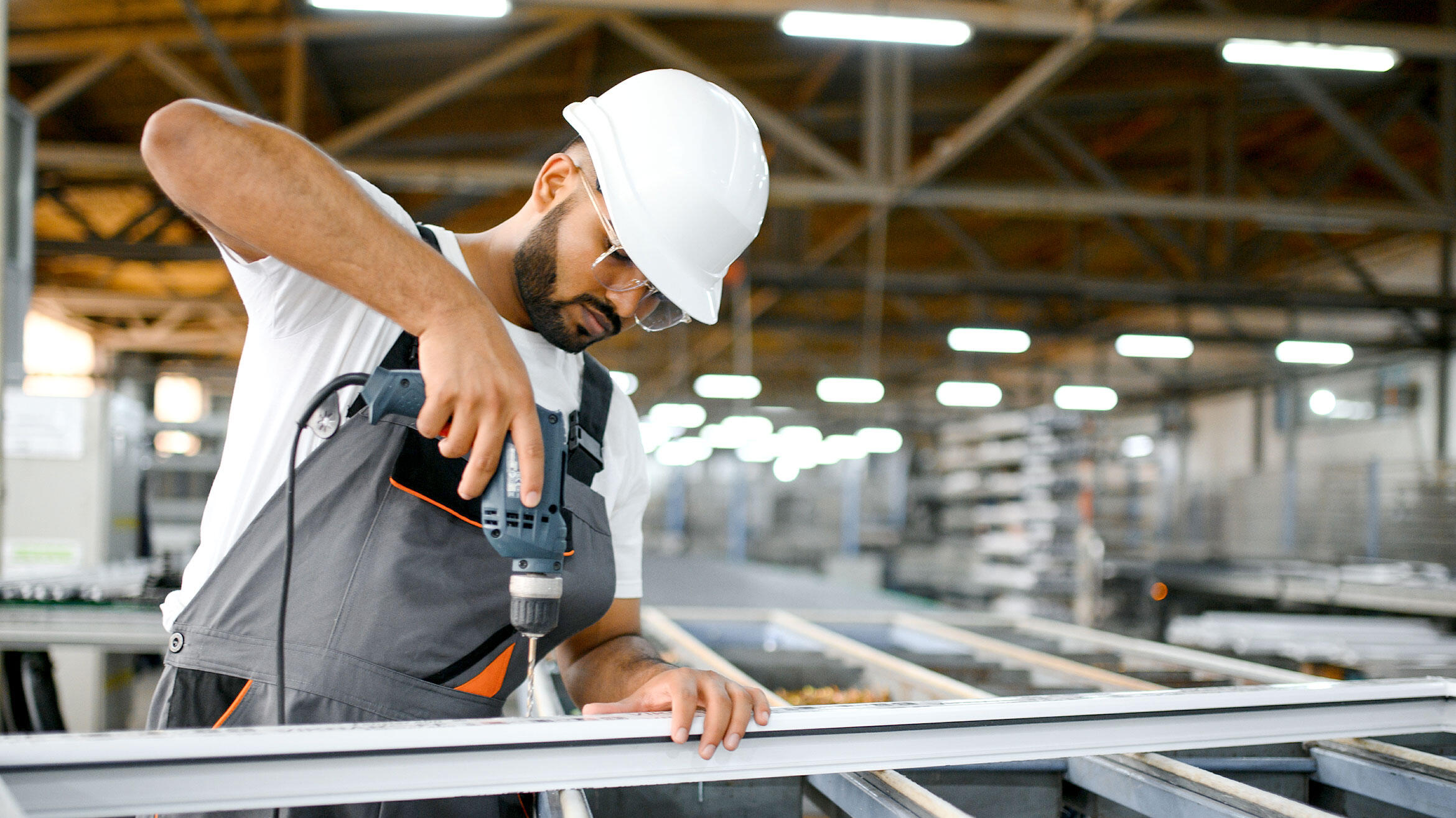 indischer Arbeiter in Arbeitskleidung und mit Schutzhelm bohrt in einen Metallrahmen