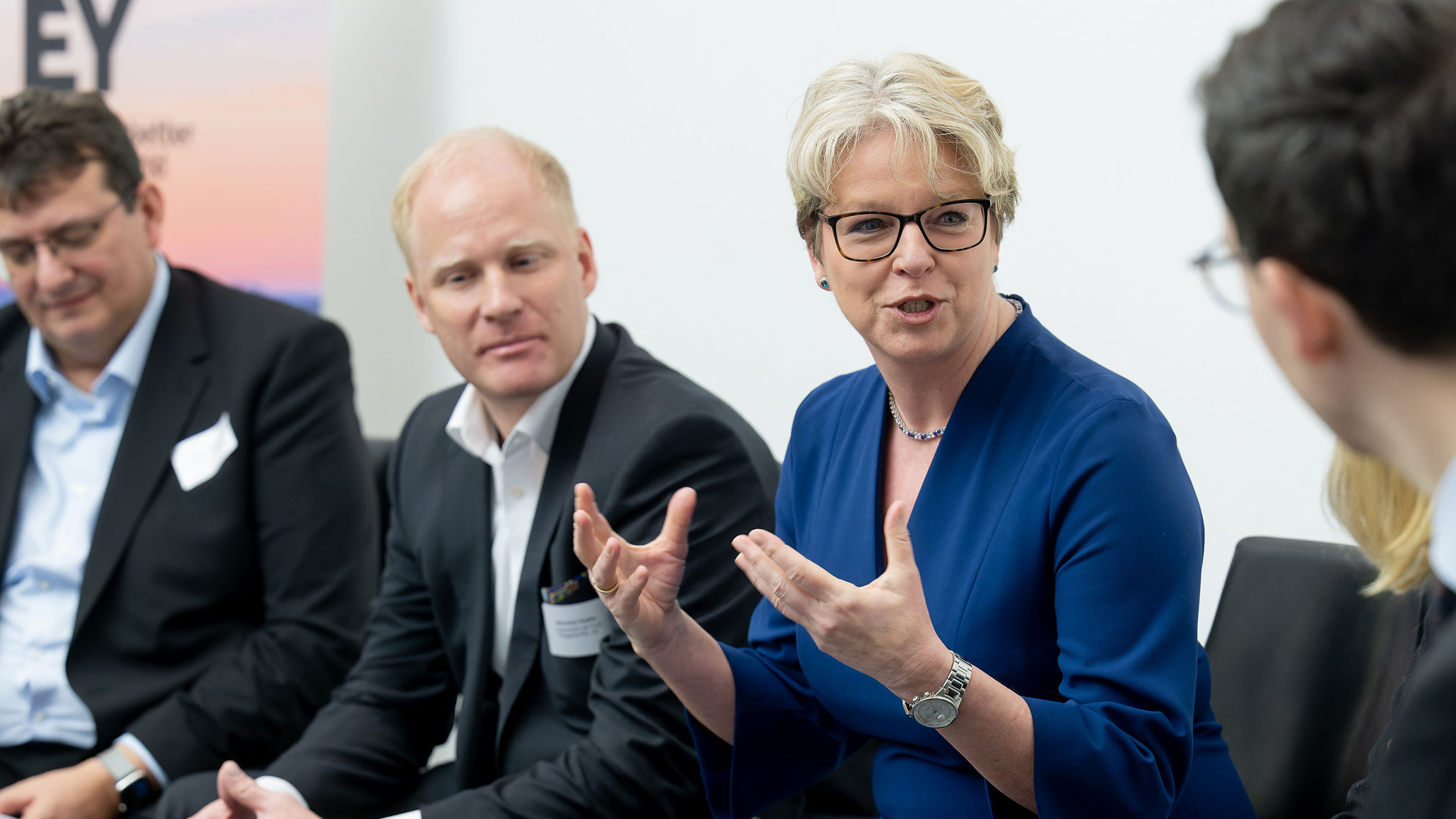 drei Personen sitzen auf dem Podium, eine Frau spricht