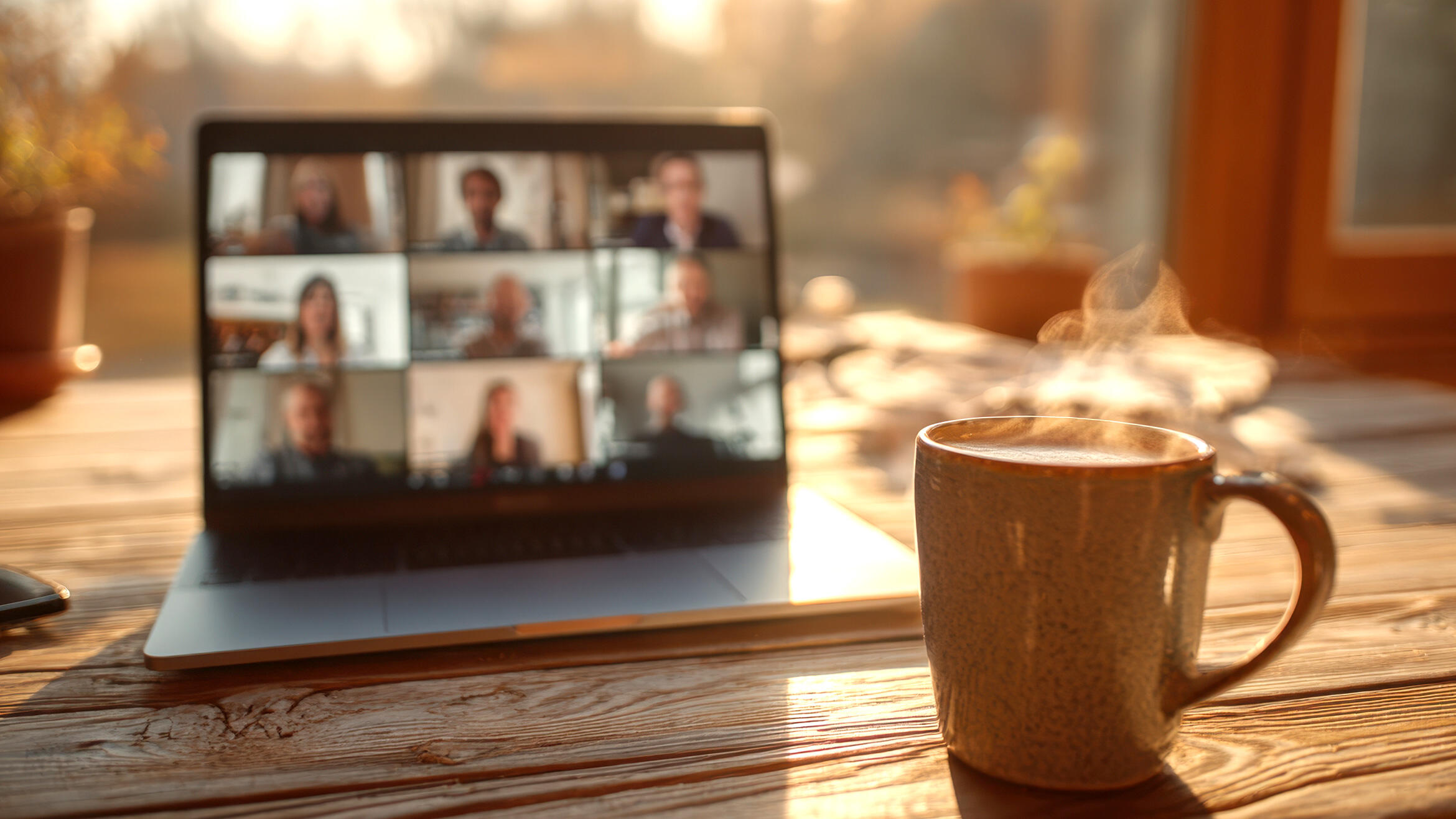 dampfende Kaffeetasse steht vor aufgeklapptem Laptop, der Bildschirm zeigt Kacheln mit sehr verschwommenen Gesichtern
