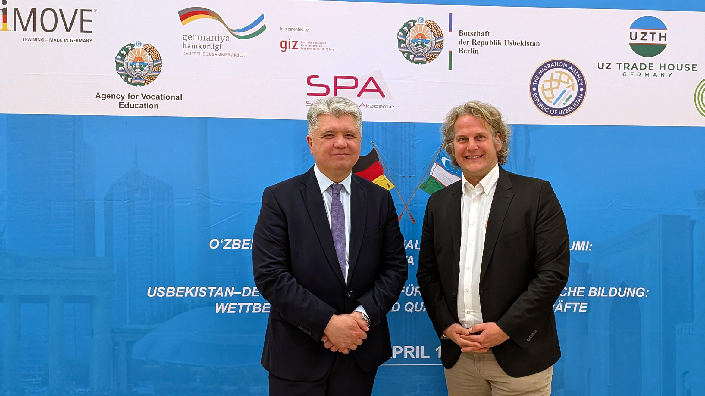 zwei Männer stehen vor einer Plakatwand der Konferenz und lächeln in die Kamera