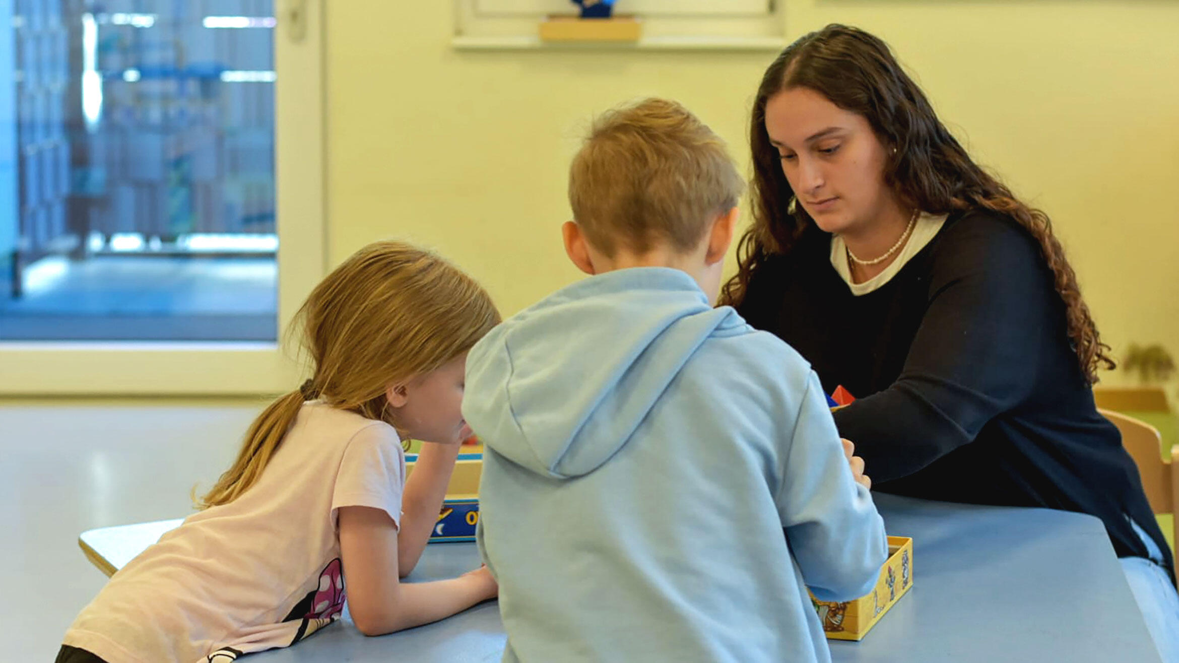 Szene in Kindertagesstätte: Junge Frau beschäftigt sich mit zwei Kindern. Die Drei sitzen an einem Tisch.