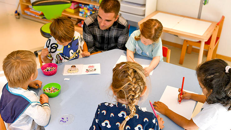 Szene in Kindertagesstätte: Pädagoge sitzt mit mehreren Mädchen und Jungen um einen Tisch herum, an dem die Kinder basteln und malen.