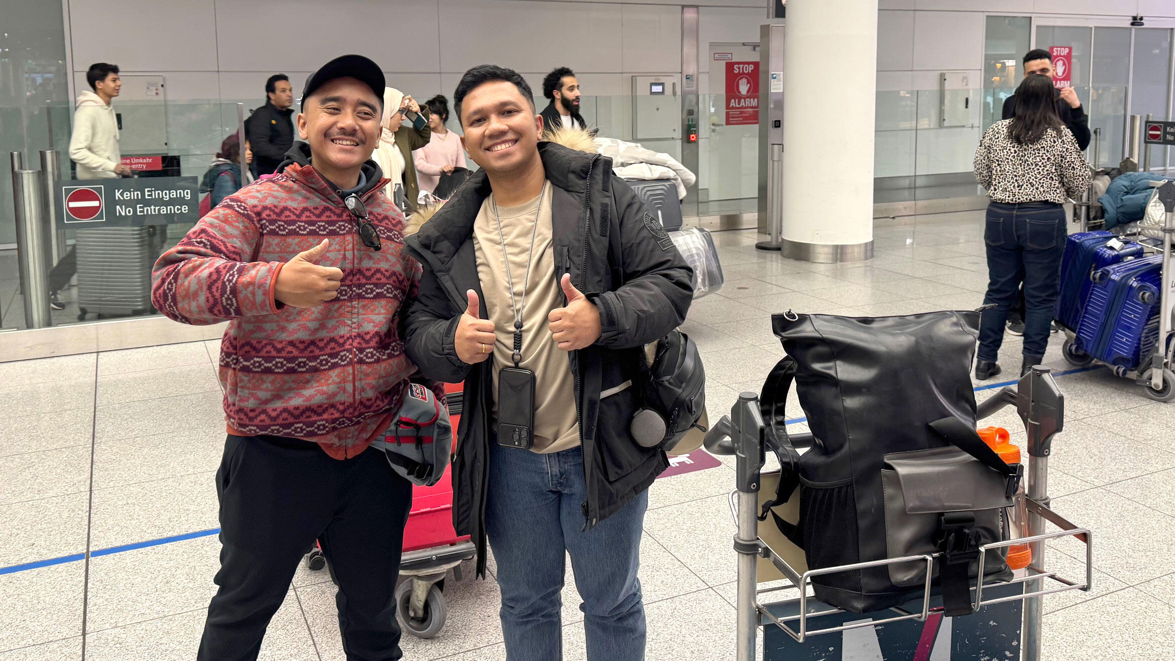 zwei Indonesier stehen mit Koffern in einer Flughafenhalle und lächeln in die Kamers, beide zeigen den Daumen hoch