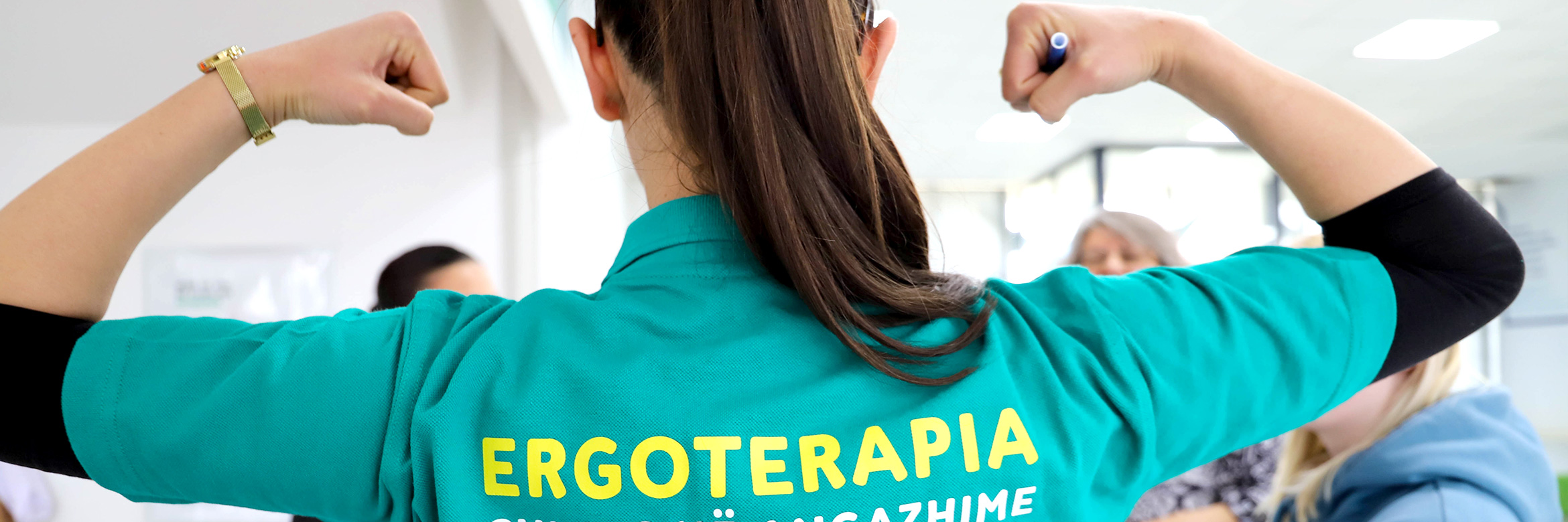 Frau von hinten fotografiert, auf ihrem Polohemd steht Ergotherapie auf ko­so­va­risch