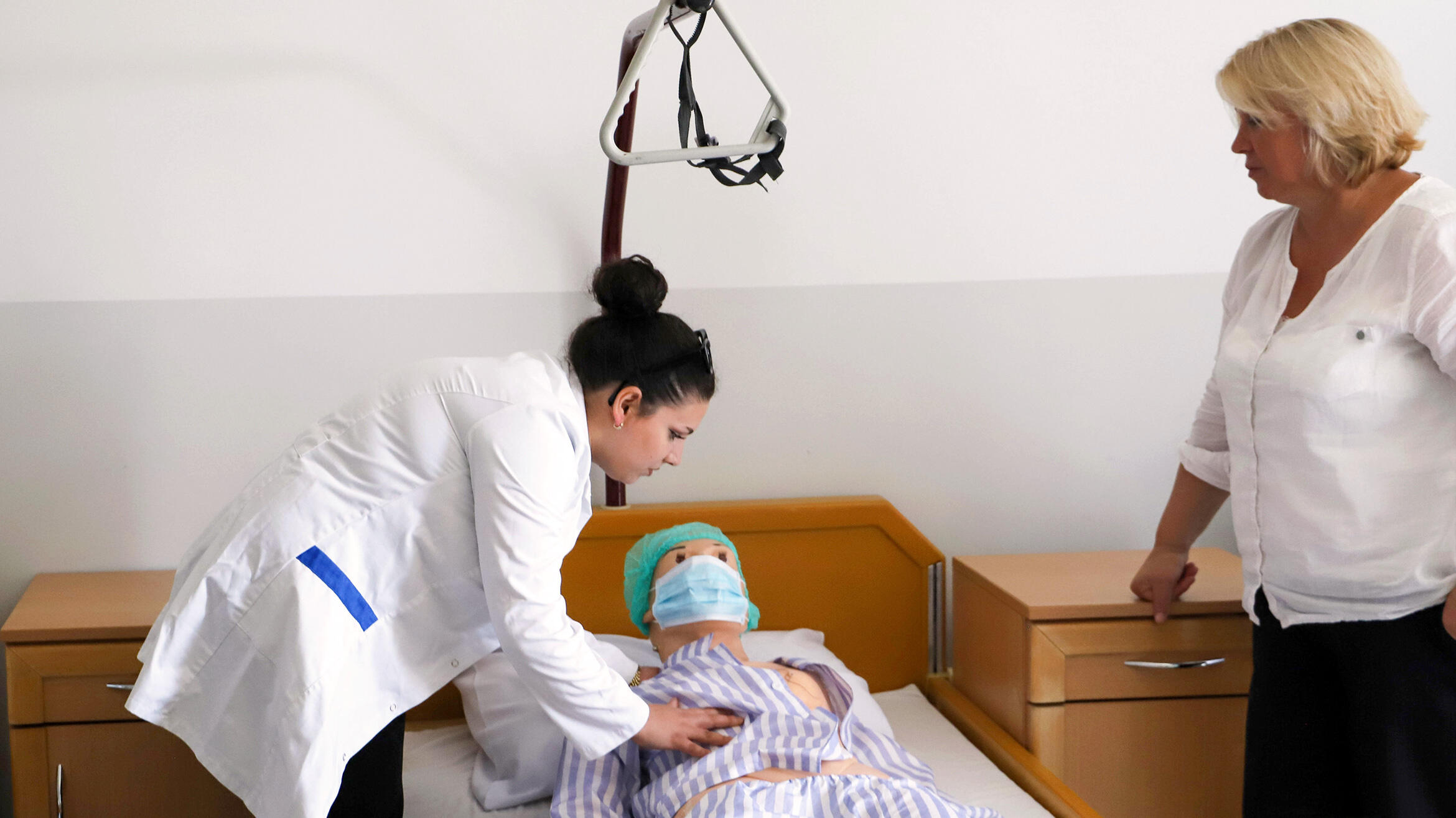 Eine junge Frau behandelt eine Puppe, die im Bett liegt. Eine weitere Frau kontrolliert die Handgriffe offenbar.