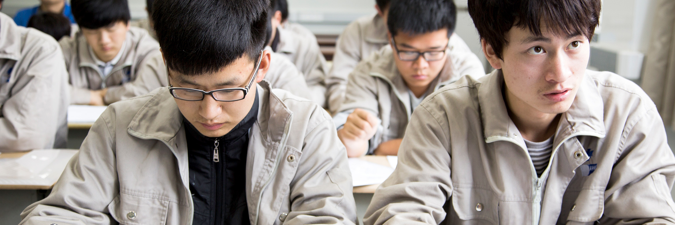 Mehrere junge Chinesen sitzen in einem Klassenraum. Sie schauen in Unterlagen auf ihren Tischen oder blicken nach vorne zu einer Lehrkraft.