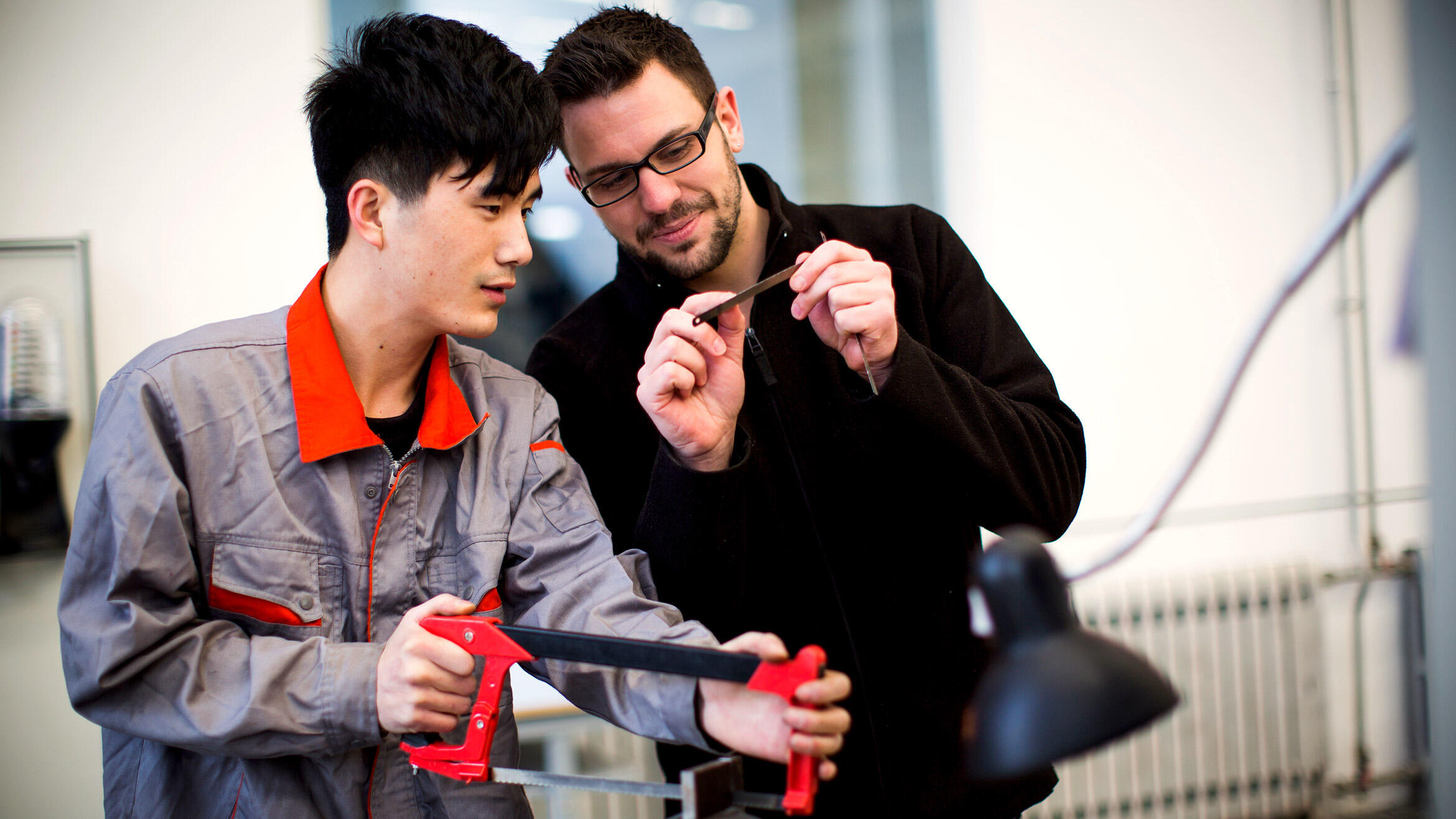 zwei Männer in Arbeitskleidung sehen sich ein Sägeblatt an, ein mutmaßlich chinesischer Mann arbeitet mit einer Handsäge