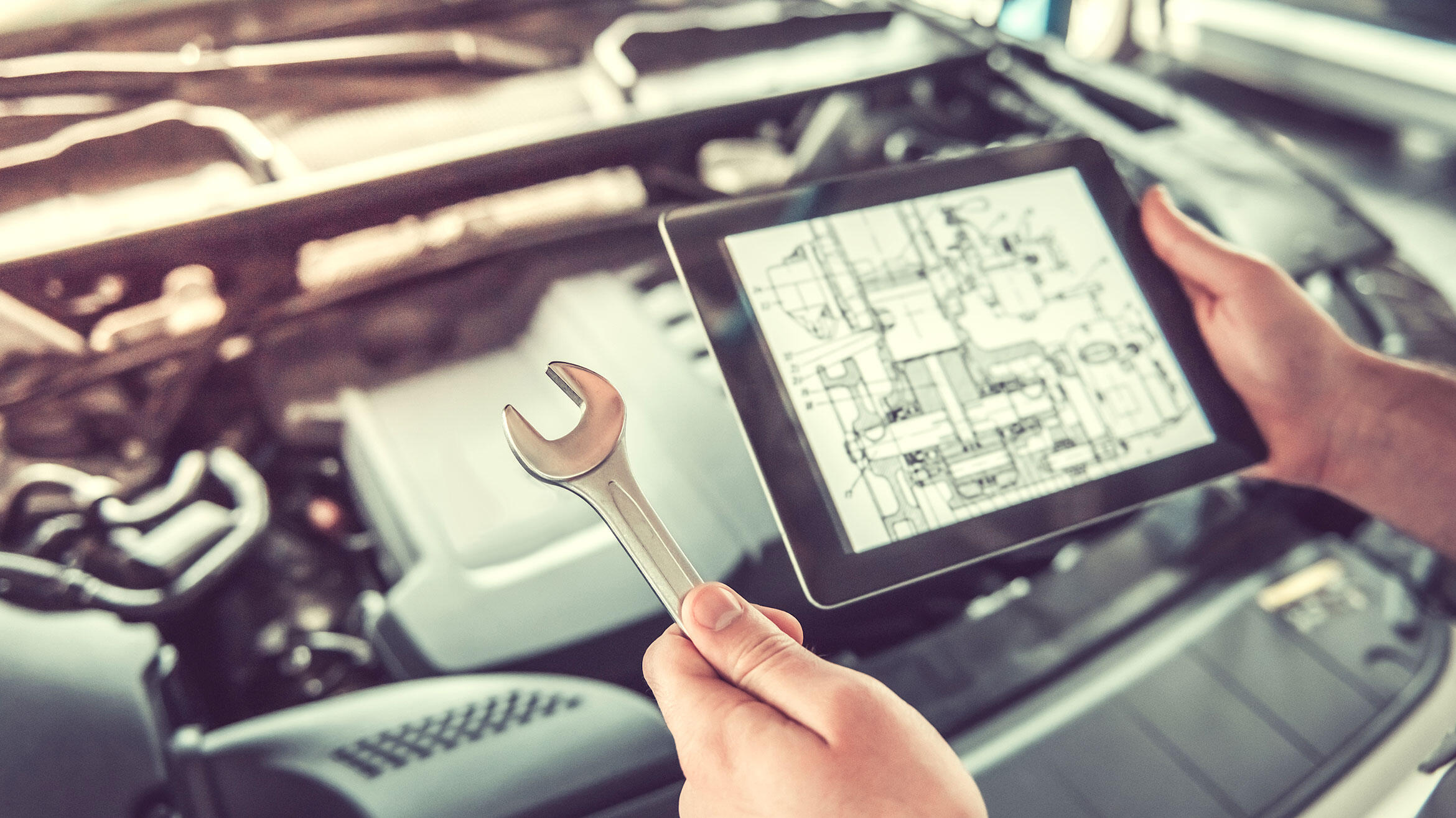 Person hält über einem Automotor in einer Hand ein Tablet  und in der anderen Hand einen Ringschlüssel