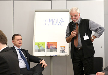 zwei Männer vor einem Flipchart mit dem Text iMOVE; ein Mann spricht in Mikrofon