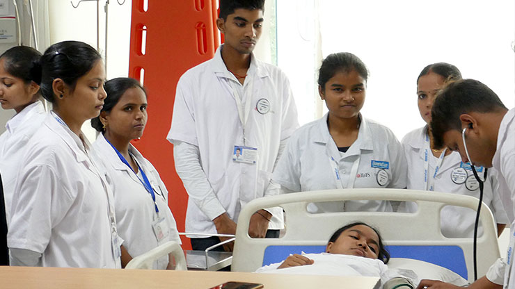 mehrere Inderinnen und Inder in Arztkitteln stehen um ein Krankenbett 