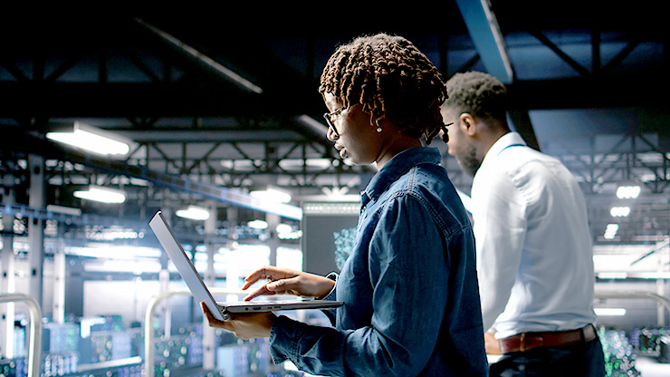 junge Frau im Vordergrund mit Laptop, dahinter junger Mann, IT Arbeitsumfeld