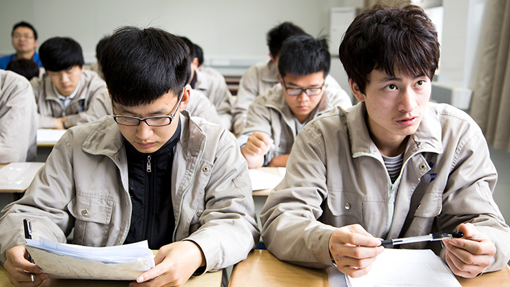 Mehrere junge Chinesen sitzen in einem Klassenraum. Sie schauen in Unterlagen auf ihren Tischen oder blicken nach vorne zu einer Lehrkraft.