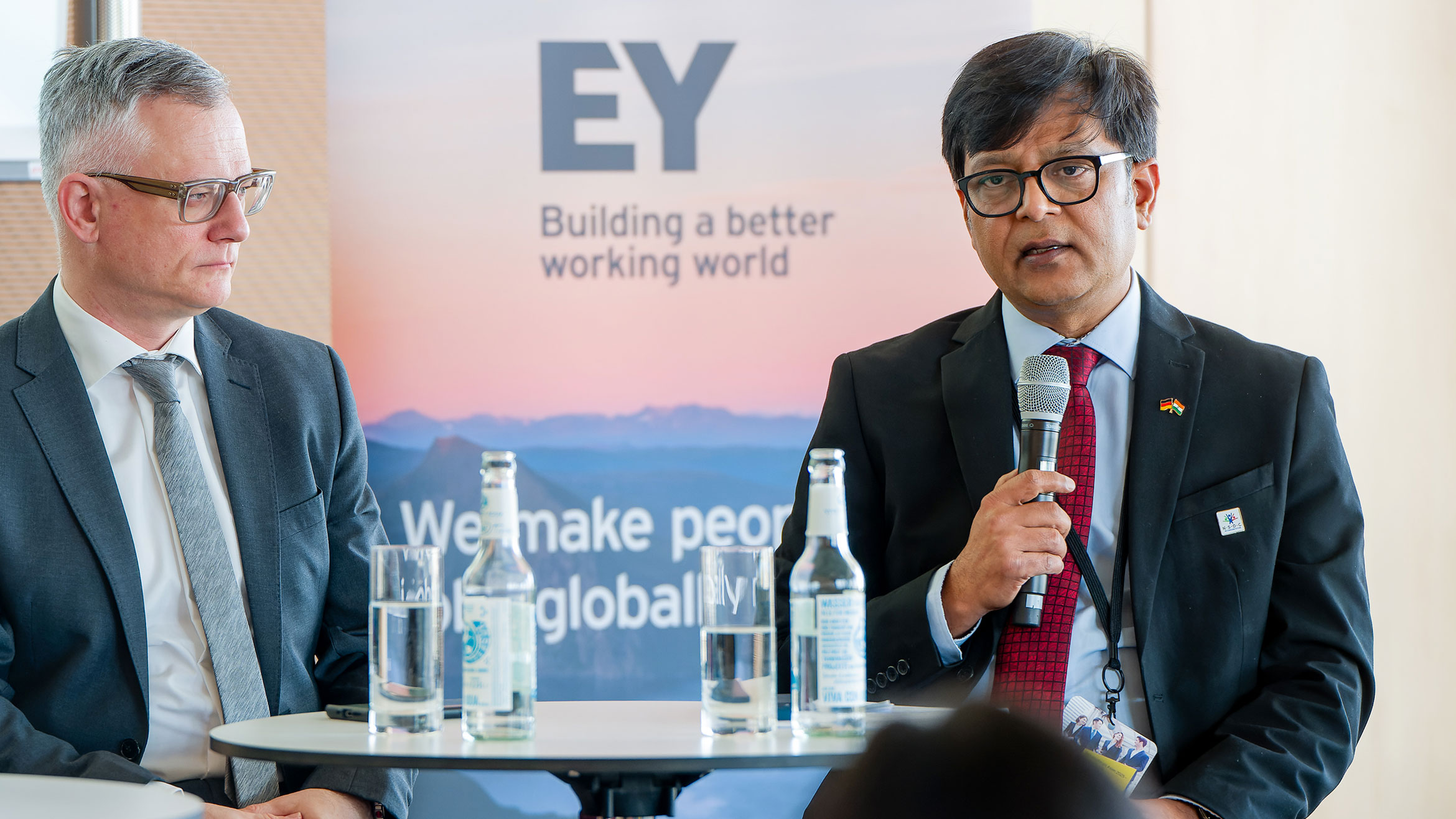 two men sit on a podium, the man on the right speaks into a microphone