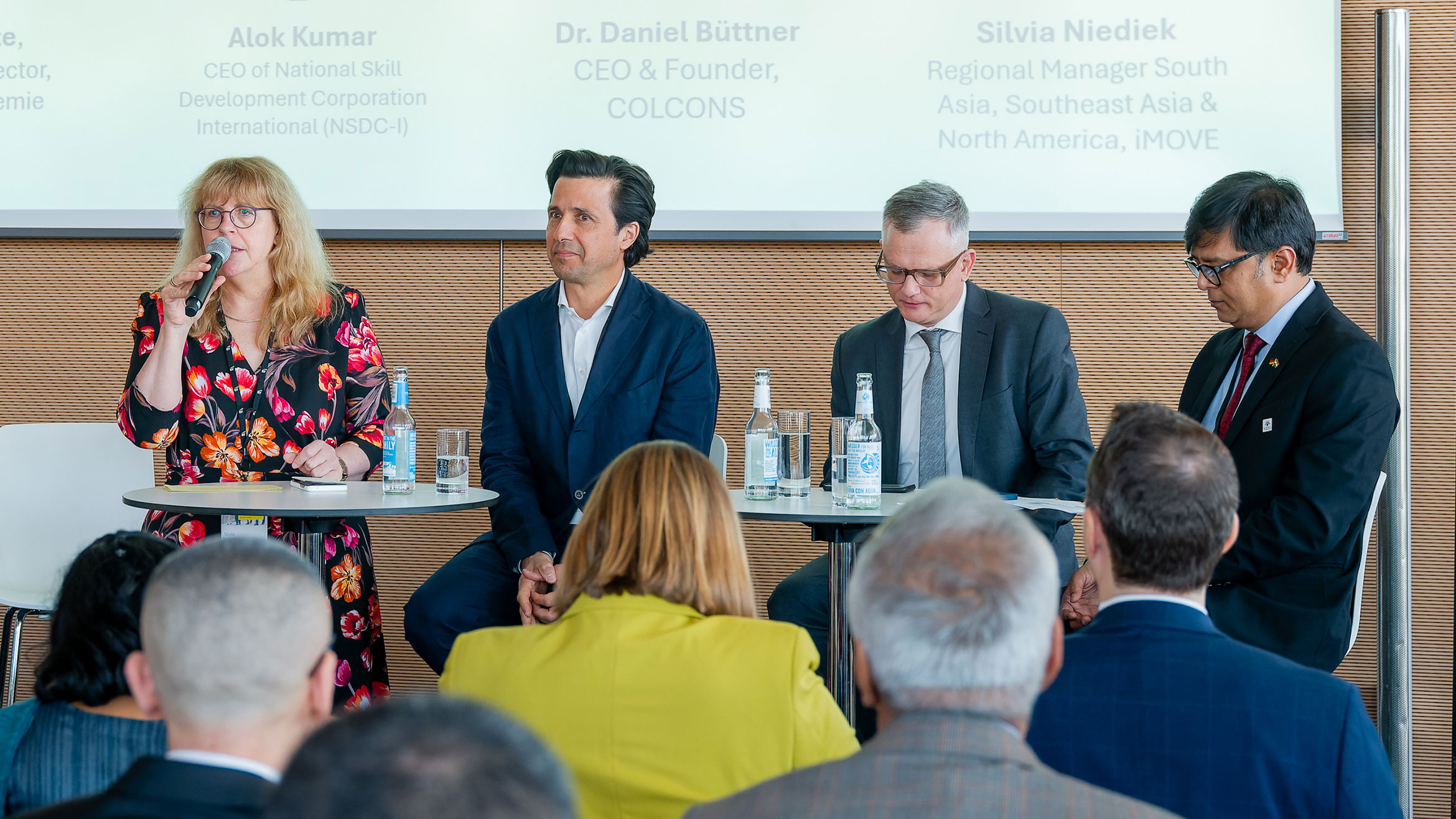 a woman and three men sit on a podium, the woman speaks into a microphone