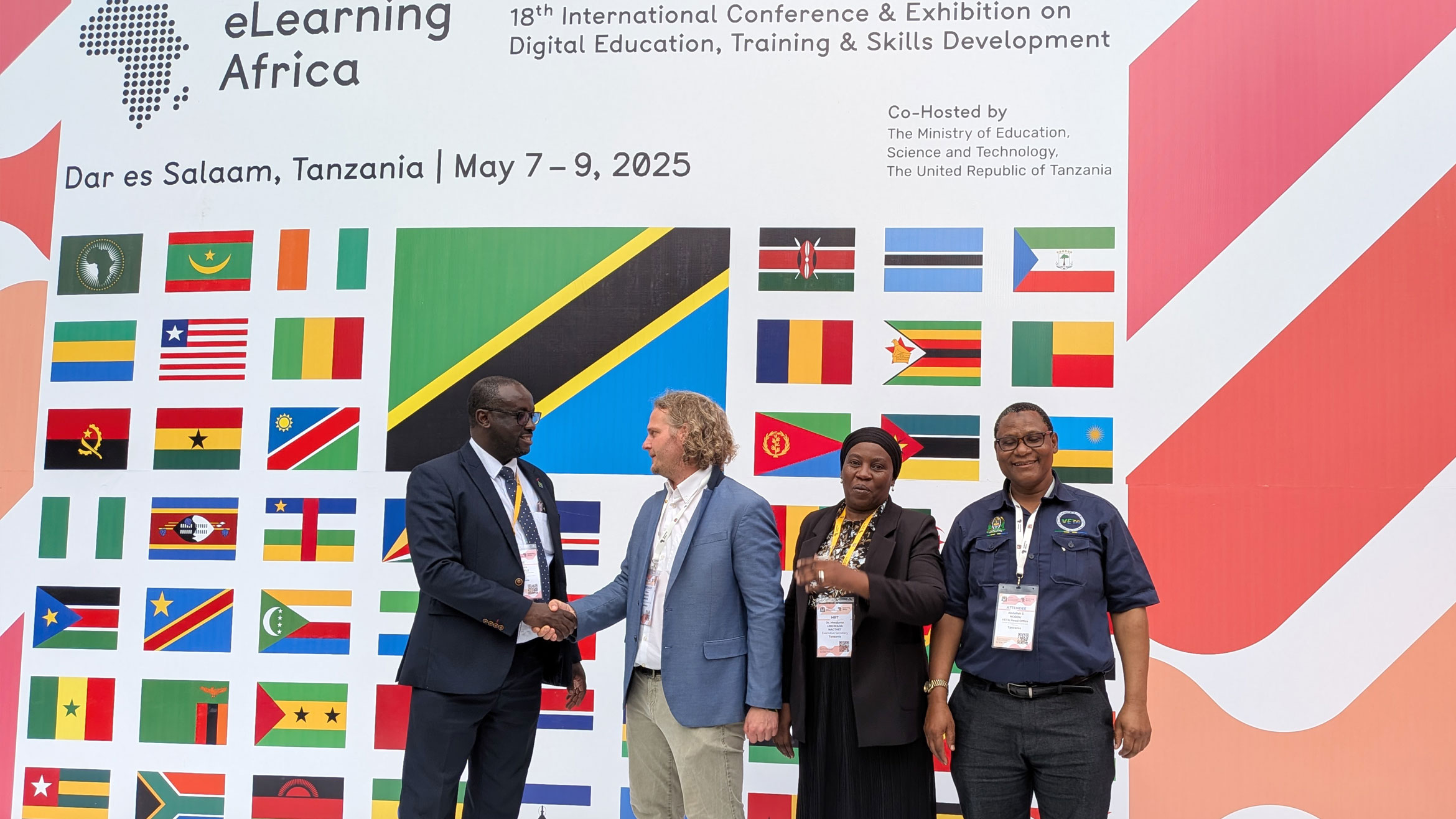 four people in a row in front of a wall with colourful national flags, two shake hands