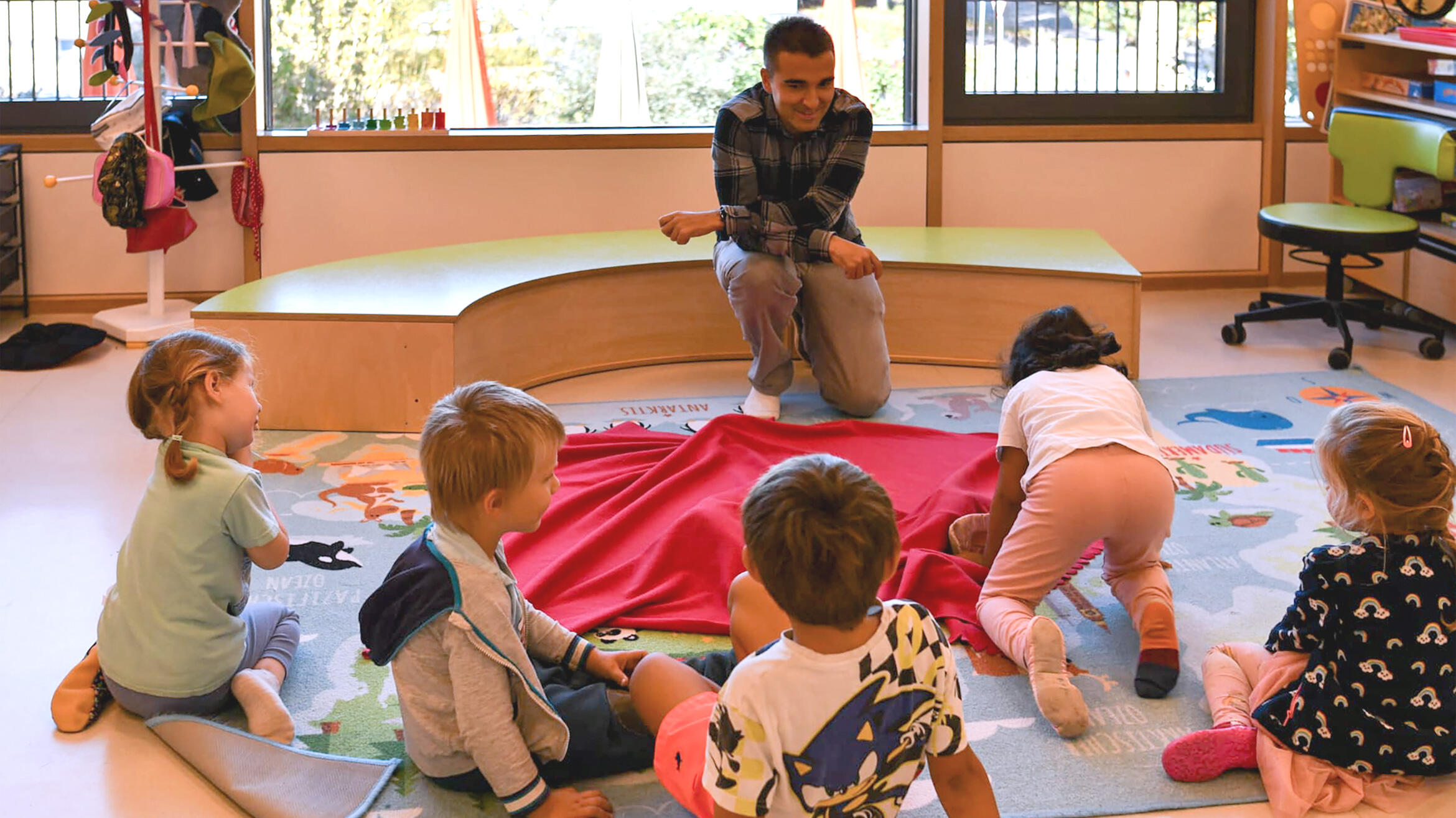 Scene in a daycare centre: Children are sitting around a blanket that is covering something. The teacher is kneeling with the children.