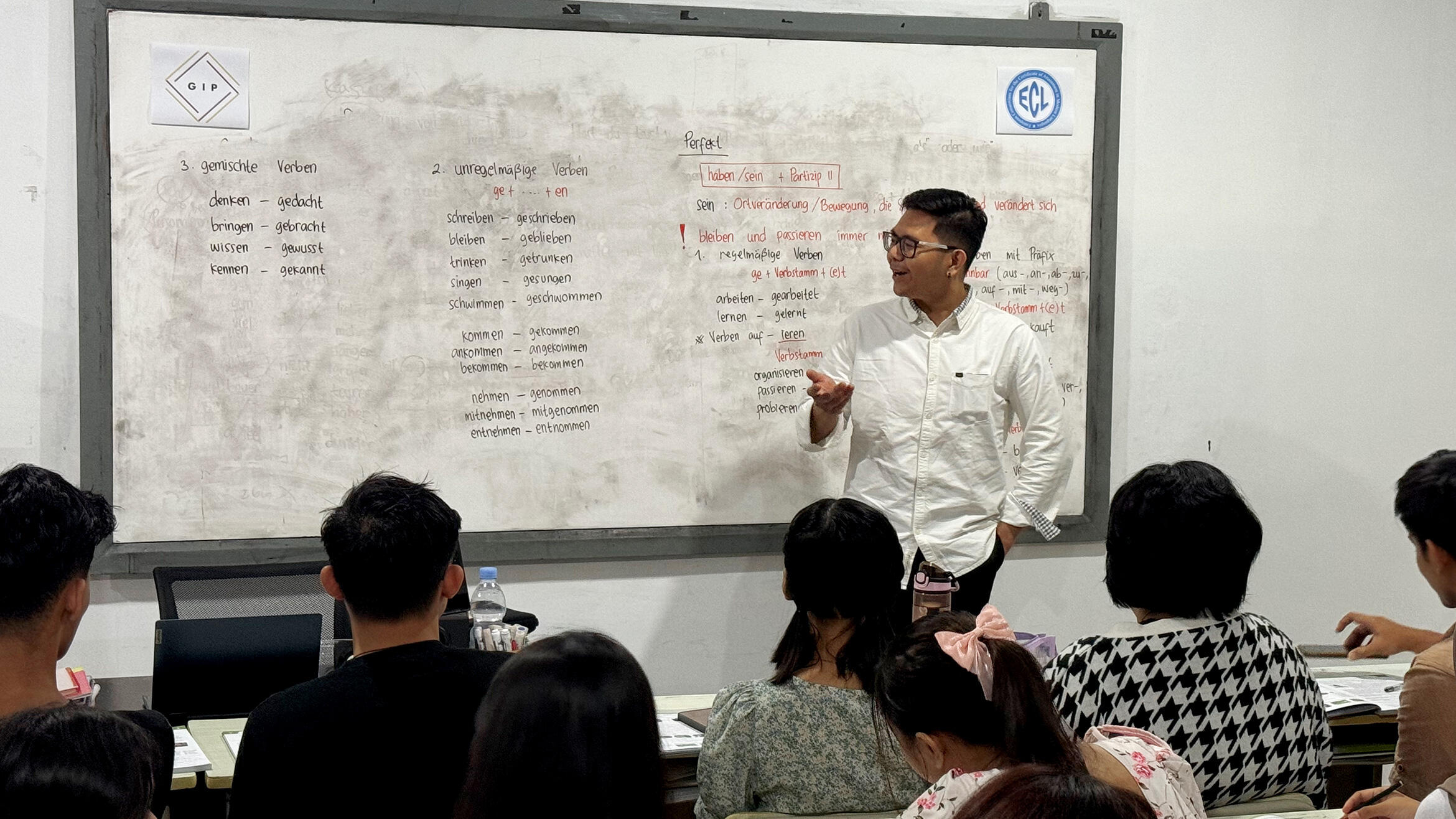 In a classroom, a man stands at the front facing the class and looks at a whiteboard behind him
