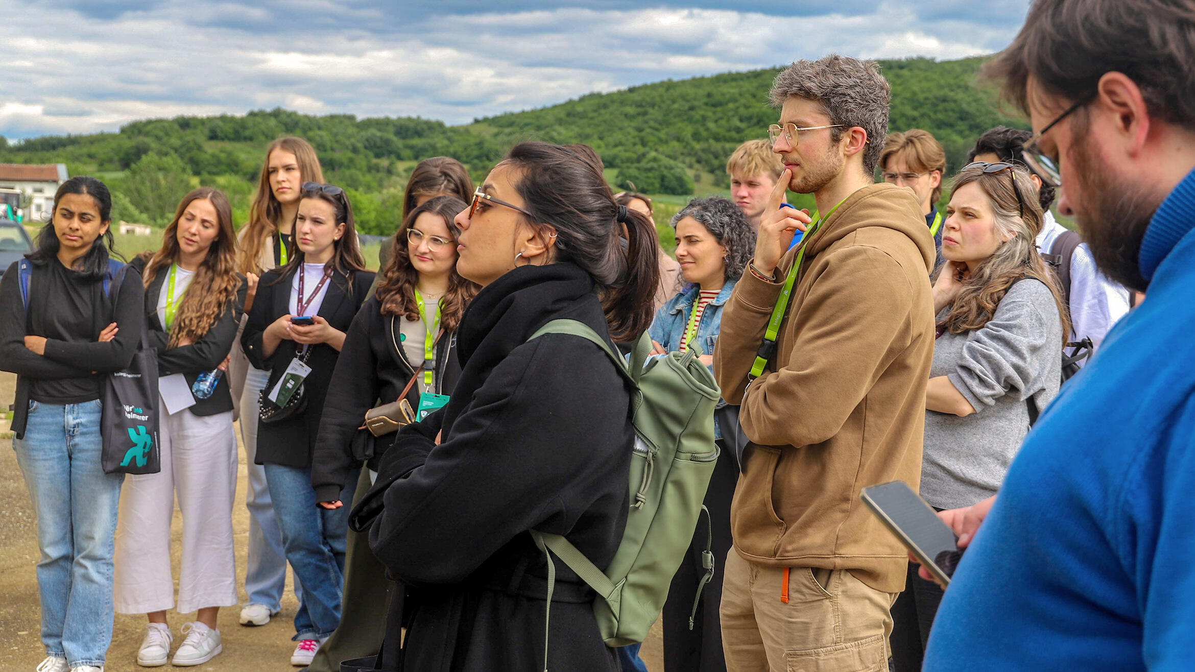 Open air: a group stands in a semicircle, all looking in the same direction and apparently listening to a person outside the picture
