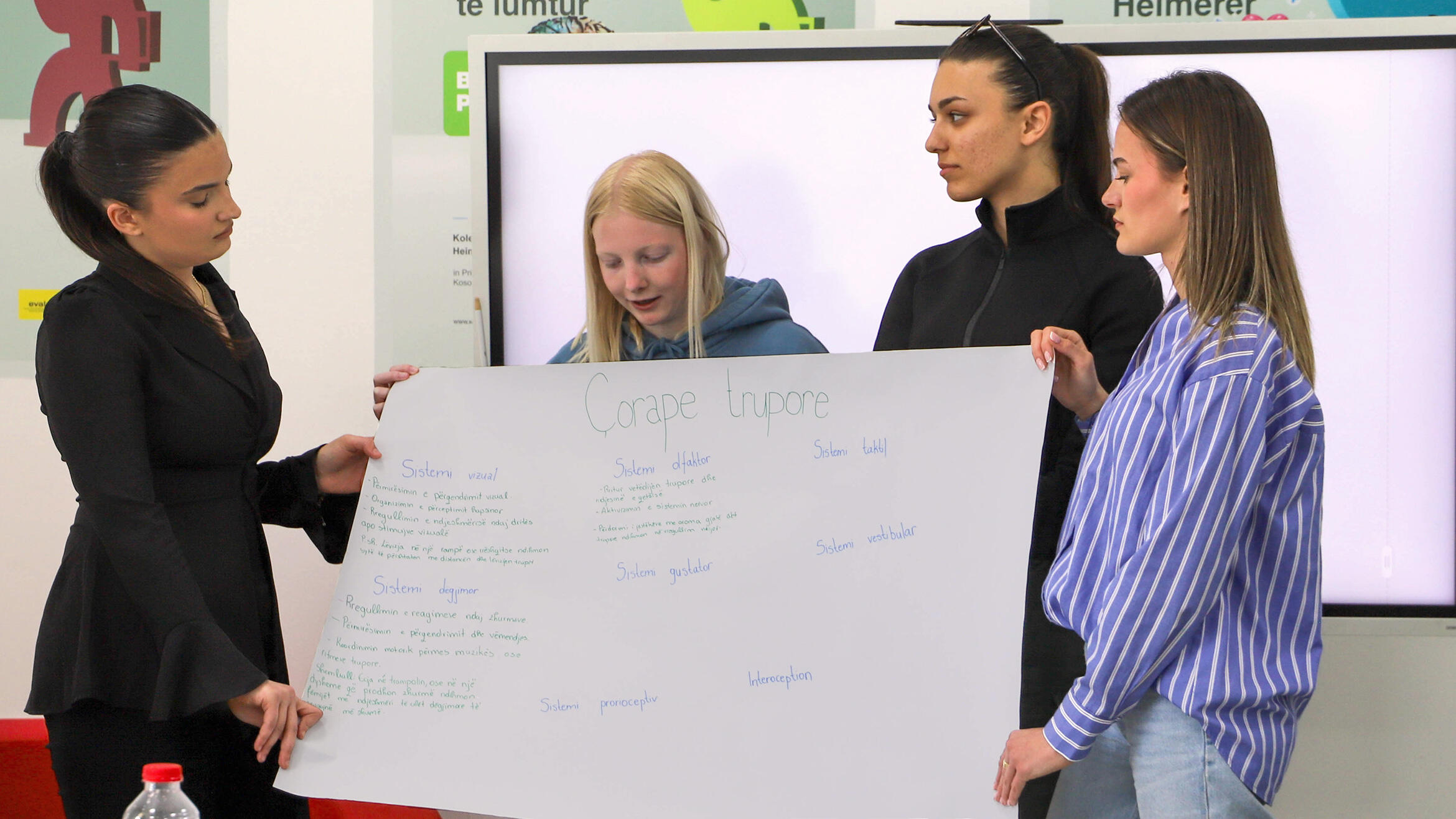 four young women holding a handwritten poster, a woman looking at the poster and speaking