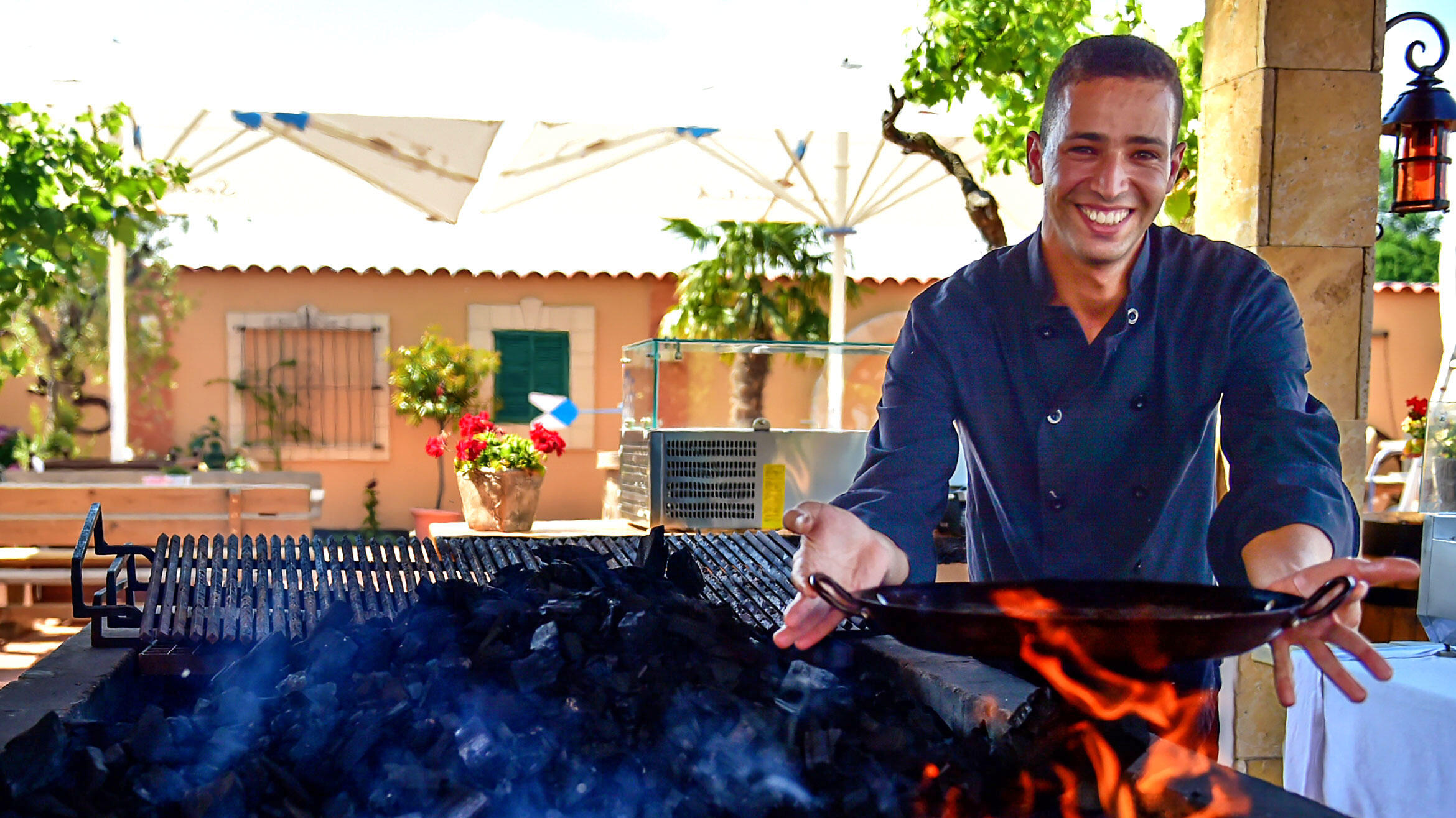 young man holds a pan over a grill fire and smiles into the camera