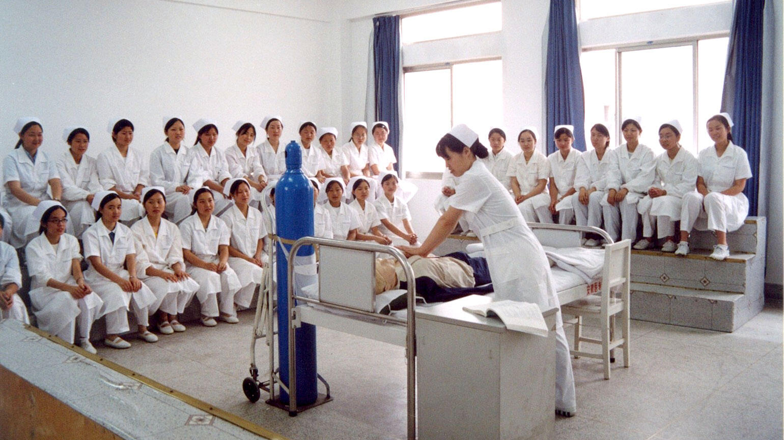 dozens of Chinese women in white coats sit in a training room, a Chinese woman treats a dummy in a hospital bed