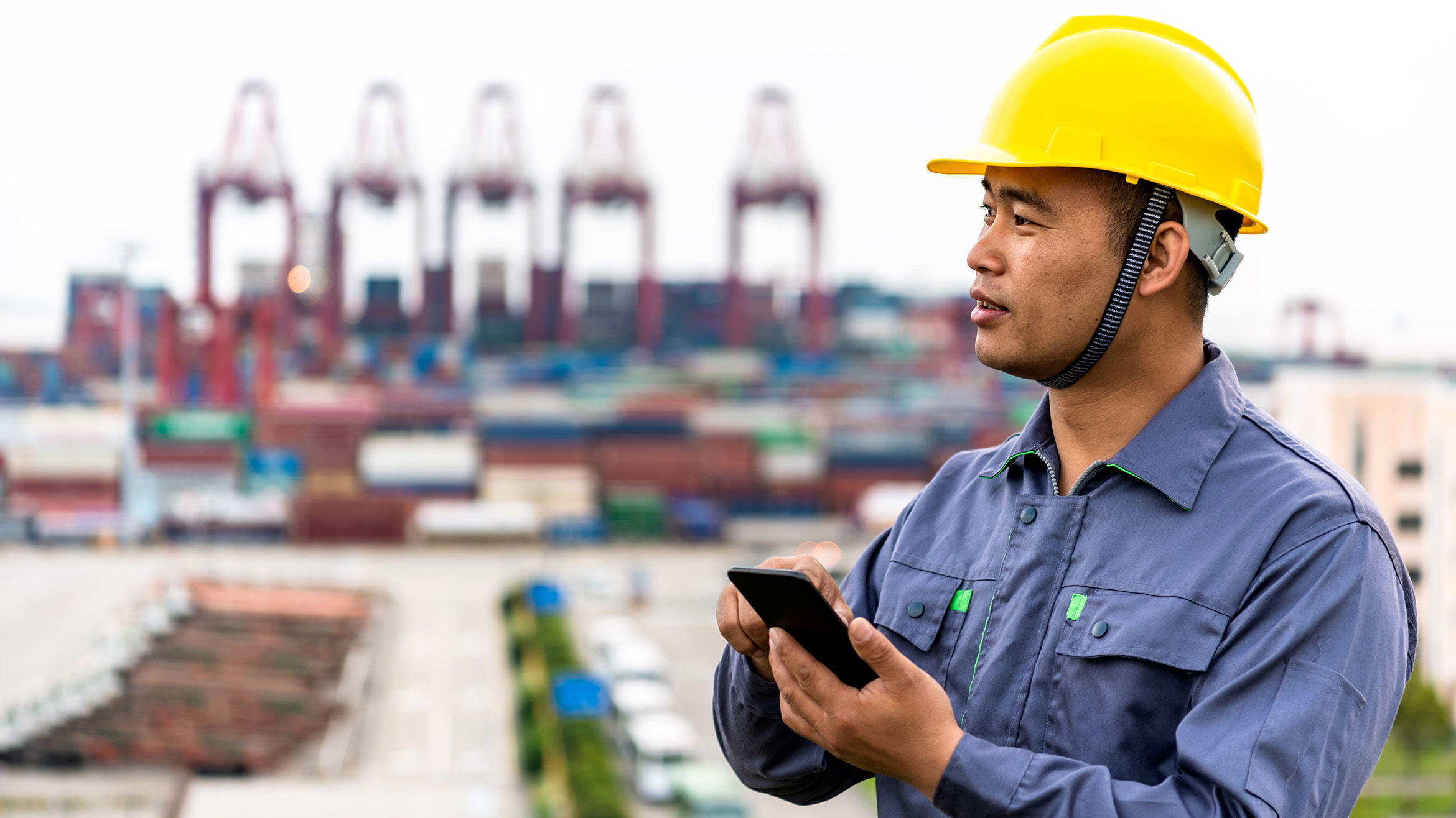 Asian man in work clothes typing on smartphone, cranes in the background