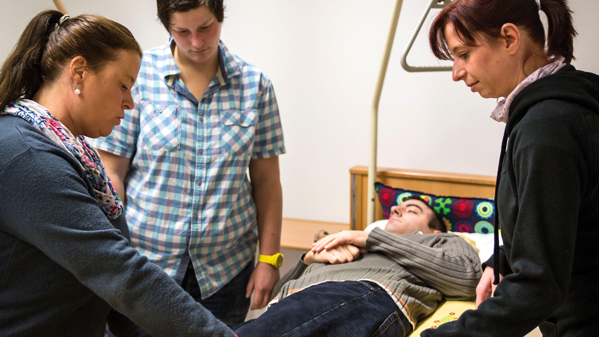 three women stand around a hospital bed in which a man lies