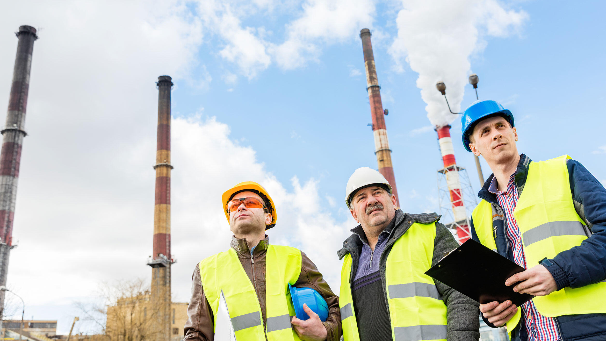 three men with safety vests and protective helmets stand in front of four chimneys