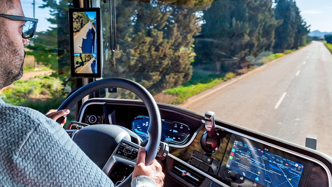 A lorry driver, photographed from the driver’s cab, is driving along a two-lane road—with one lane in each direction—which winds through the countryside