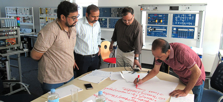 four Indien men stand around a table and talk about a chart