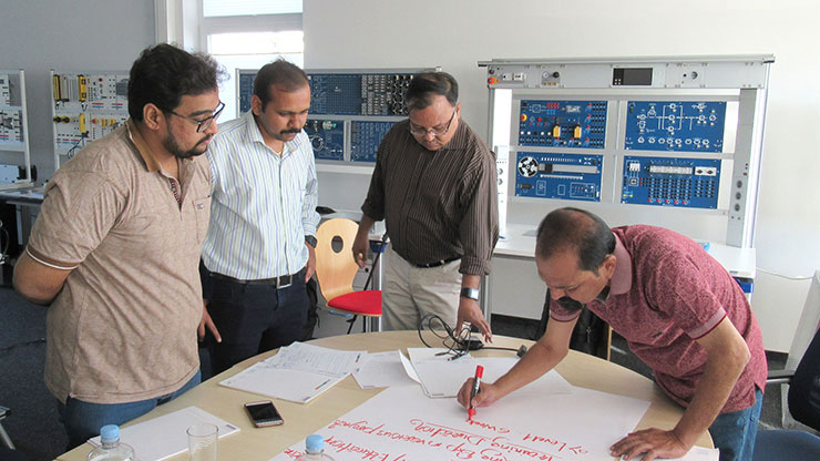 four Indien men stand around a table and talk about a chart