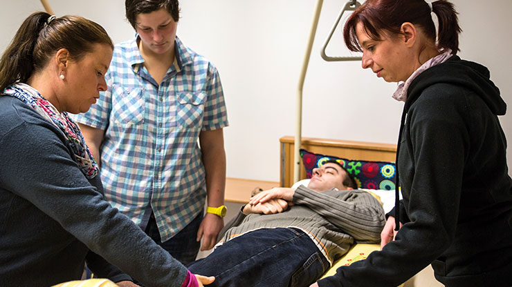 three women stand around a hospital bed in which a man lies